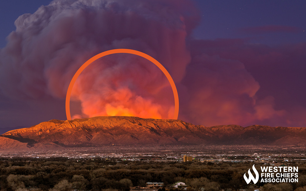 An orange mesa overlooks a town at sunset. The glow of fire behind the mesa is highlighted with an orange semi-circle.